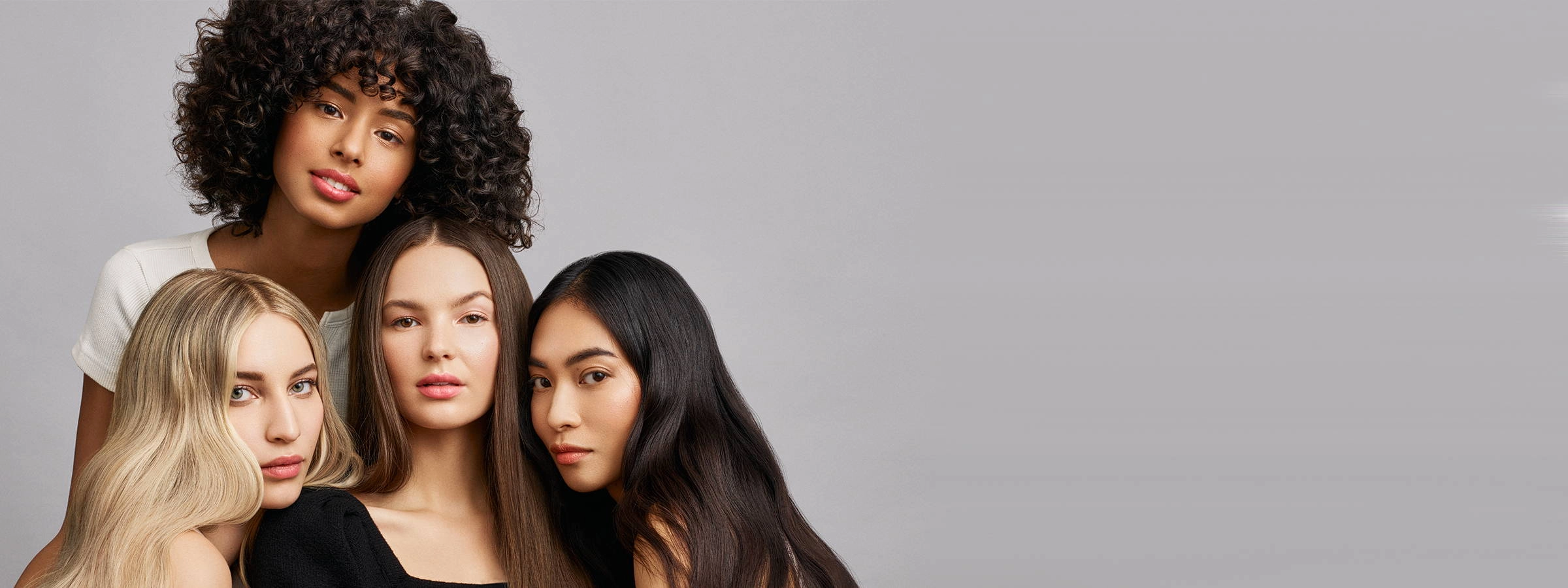 Four women with curly, light brown, straight and black hair posing close together against a grey backdrop